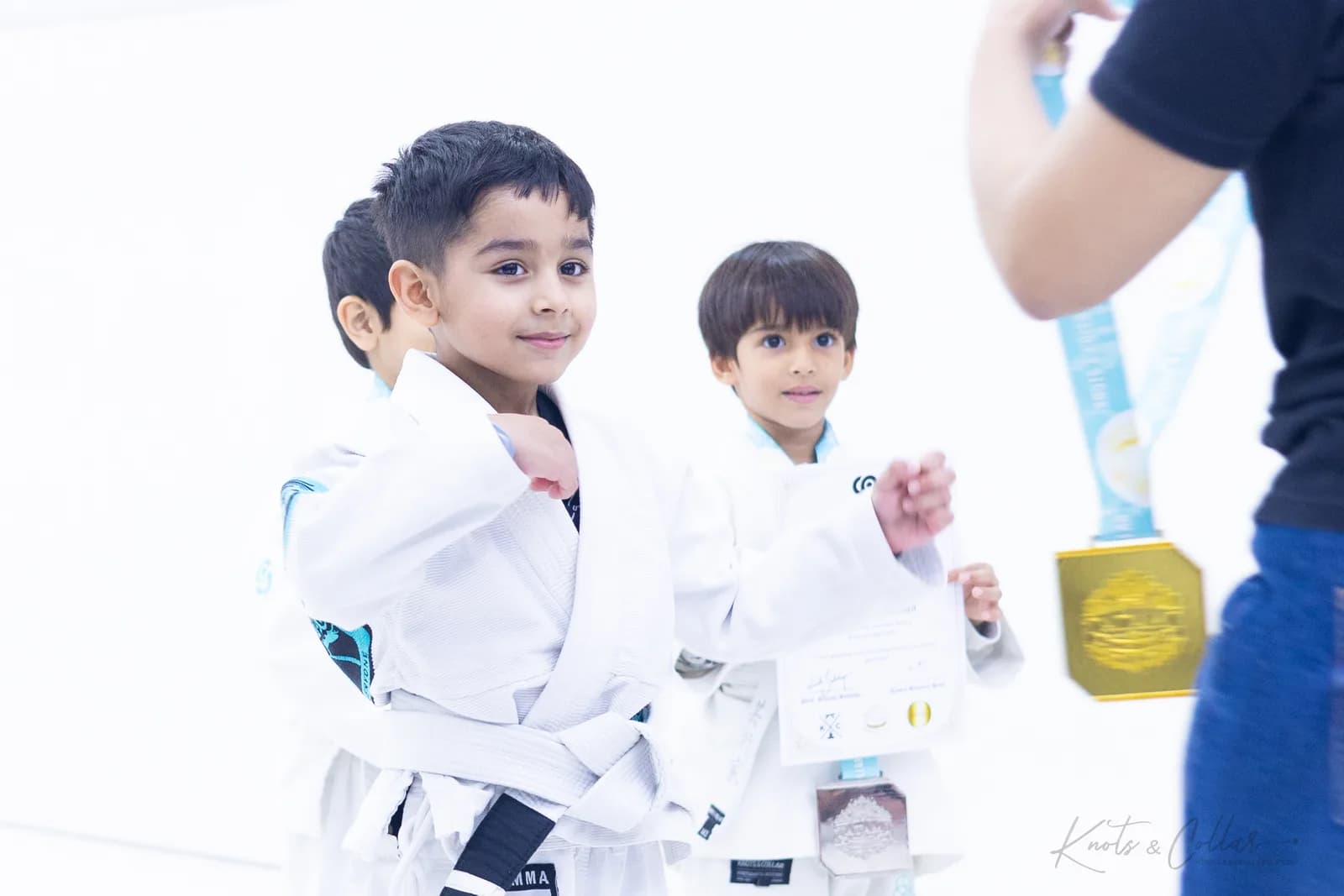 Two young Knots & Collar students in white gis proudly holding their Brazilian Jiu-Jitsu grading certificate and medal at the kids BJJ class in Defence Colony, New Delhi